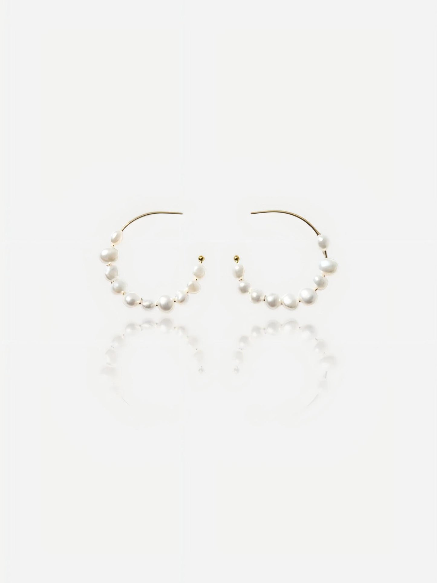 Top-down view of Pearltide Isla organic hoop pearl jewelry earrings featuring dainty gold C-hoops threaded with petite white baroque pearls on a textured beige backdrop, elegant minimalist resort jewelry.