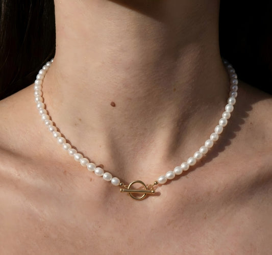 A close-up of a woman's neck wearing a classic white pearl necklace. The gold toggle clasp is worn in the front, centered on the collarbone, highlighting a vintage French aesthetic.