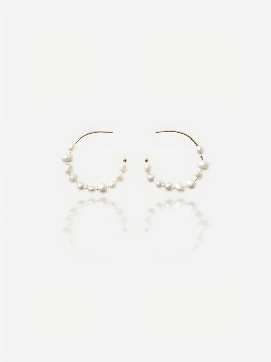 Top-down view of Pearltide Isla organic hoop pearl jewelry earrings featuring dainty gold C-hoops threaded with petite white baroque pearls on a textured beige backdrop, elegant minimalist resort jewelry.