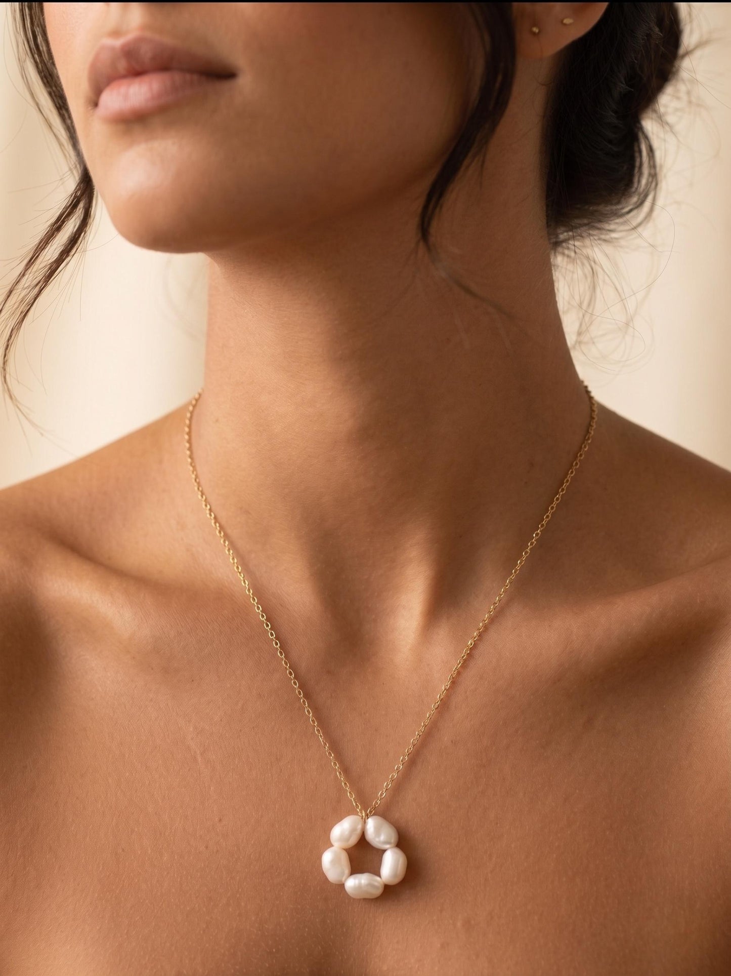 Close-up profile of a model with glowing tan skin wearing a delicate gold chain with a single large, naturally textured, irregular baroque pearl pendant against a white background, embodying minimalist elegance for bridal or everyday style.