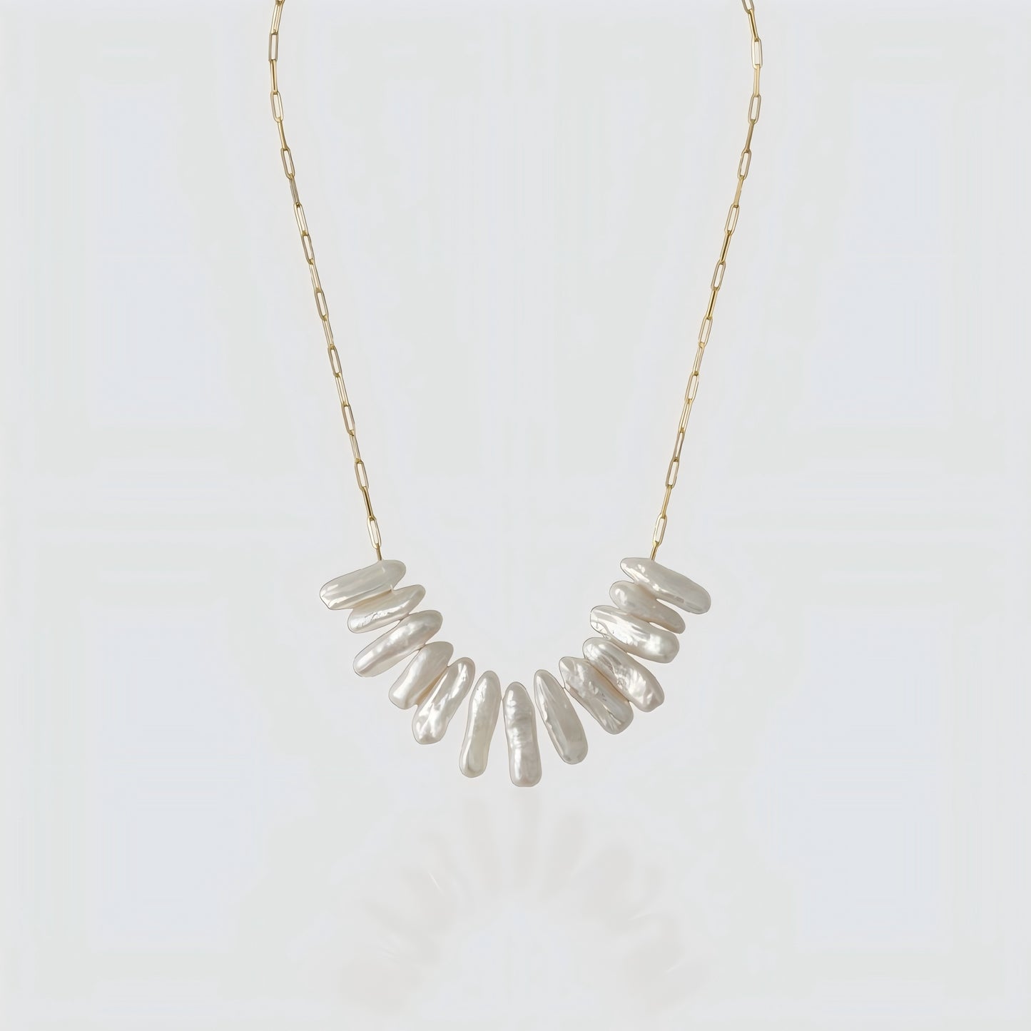 A studio shot of a statement necklace featuring a cluster of long, iridescent stick pearls on a gold paperclip chain, set against a grey background.