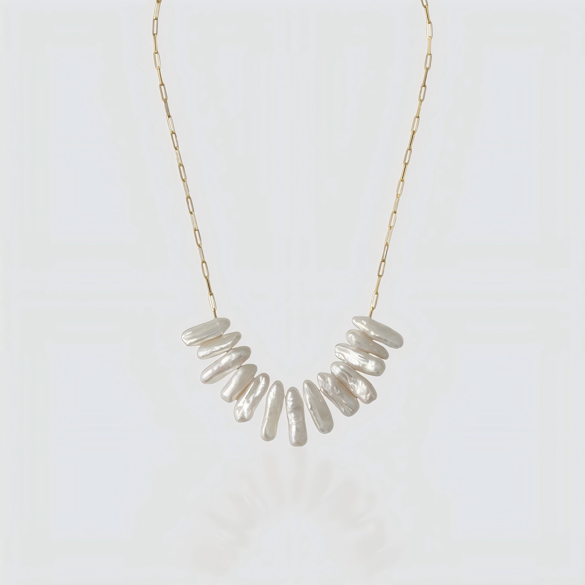 A studio shot of a statement necklace featuring a cluster of long, iridescent stick pearls on a gold paperclip chain, set against a grey background.