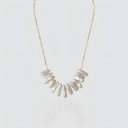 A studio shot of a statement necklace featuring a cluster of long, iridescent stick pearls on a gold paperclip chain, set against a grey background.