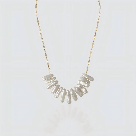 A studio shot of a statement necklace featuring a cluster of long, iridescent stick pearls on a gold paperclip chain, set against a grey background.