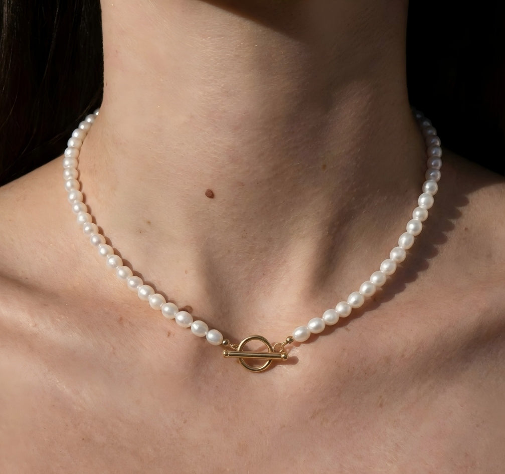 A close-up of a woman's neck wearing a classic white pearl necklace. The gold toggle clasp is worn in the front, centered on the collarbone, highlighting a vintage French aesthetic.