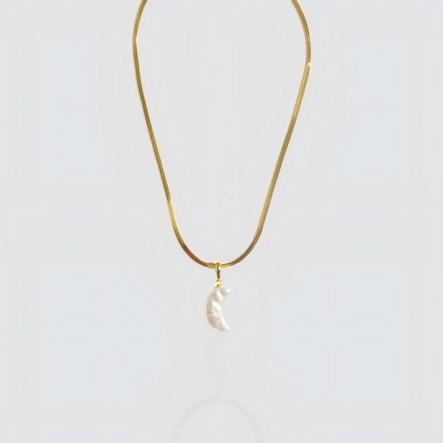 Studio shot of a white pearl crescent moon pendant attached to a flat gold snake chain on a grey background.