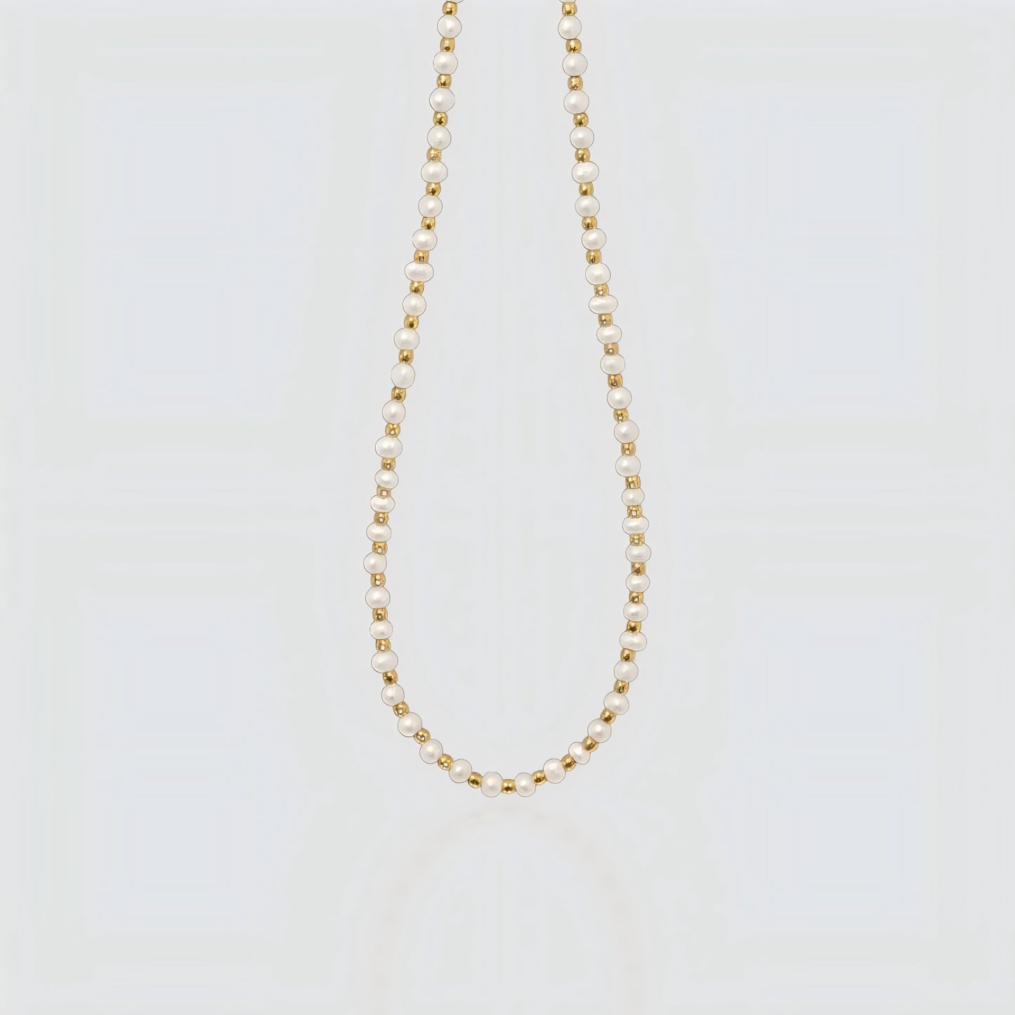 Studio product shot of a delicate necklace featuring alternating small white freshwater pearls and gold beads on a light grey background.
