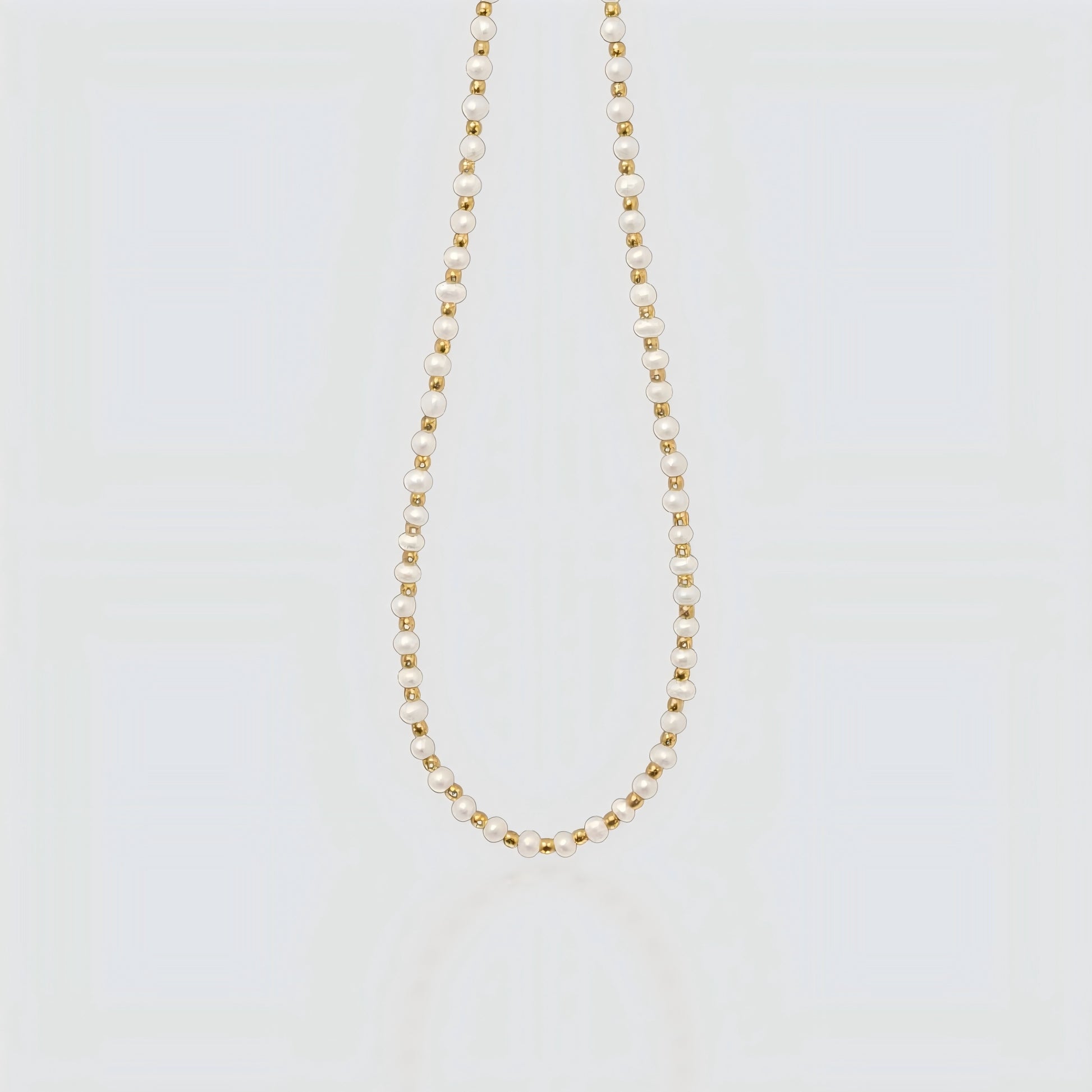 Studio product shot of a delicate necklace featuring alternating small white freshwater pearls and gold beads on a light grey background.