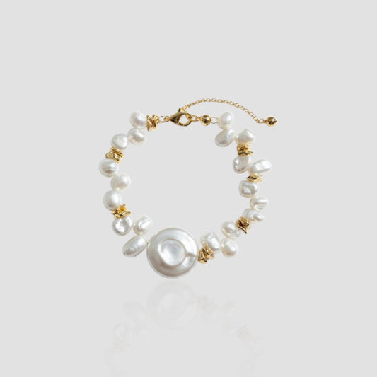 Studio shot of a bracelet featuring a large flat white coin pearl in the center, surrounded by baroque pearls and gold spacer beads on a light grey background.