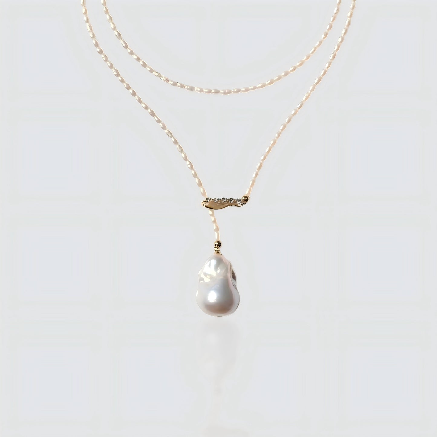 A studio shot of a dainty seed pearl necklace with a gold crystal bar and a baroque pearl drop, isolated on a white background.