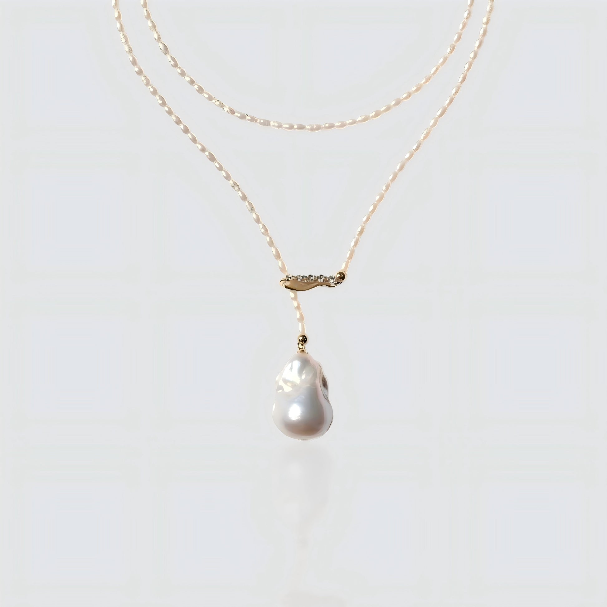 A studio shot of a dainty seed pearl necklace with a gold crystal bar and a baroque pearl drop, isolated on a white background.