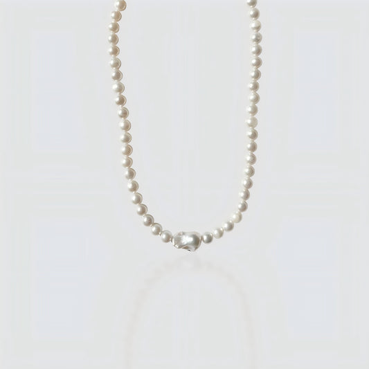 Studio shot of a large pearl strand necklace culminating in a massive, metallic-luster baroque Edison pearl centerpiece on a textured stone surface.