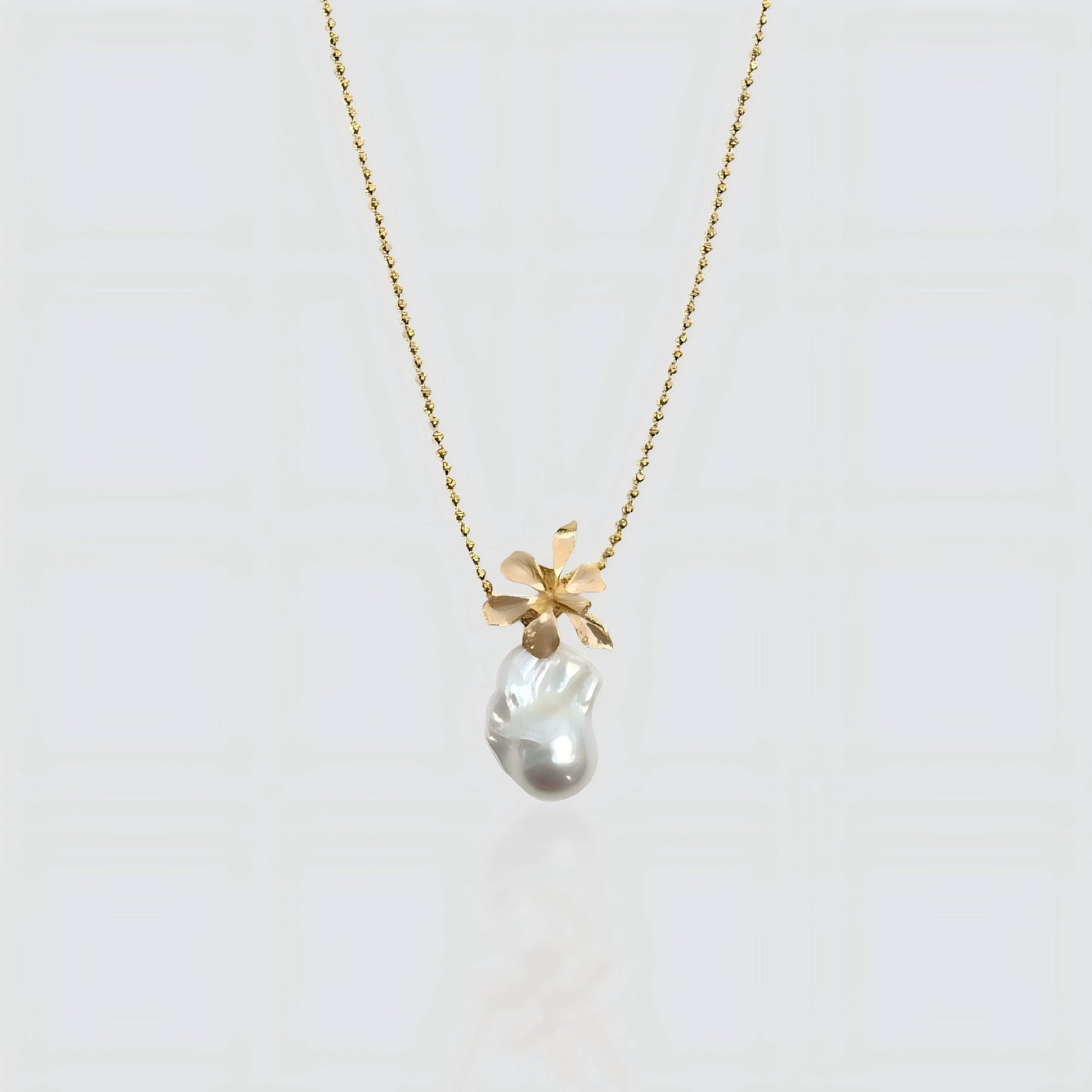 A white freshwater baroque pearl pendant topped with a sculpted gold flower design, hanging on a gold beaded satellite chain against a white background.