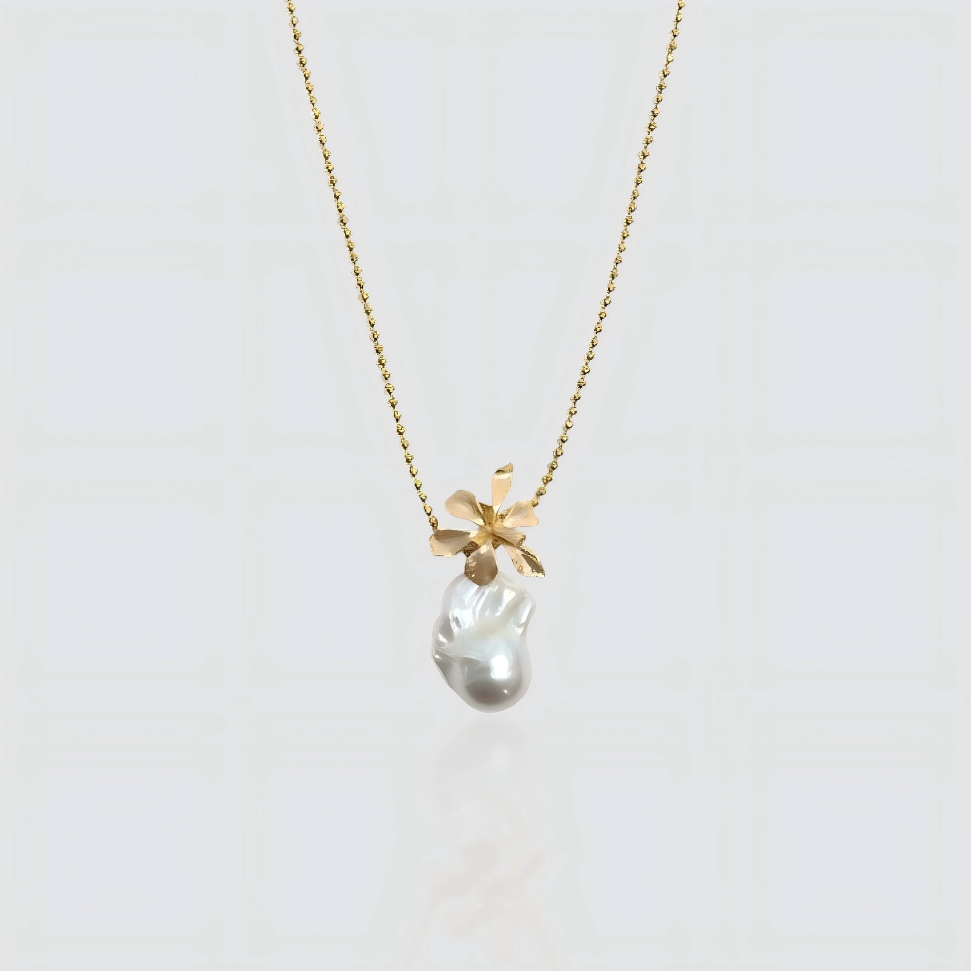 A white freshwater baroque pearl pendant topped with a sculpted gold flower design, hanging on a gold beaded satellite chain against a white background.
