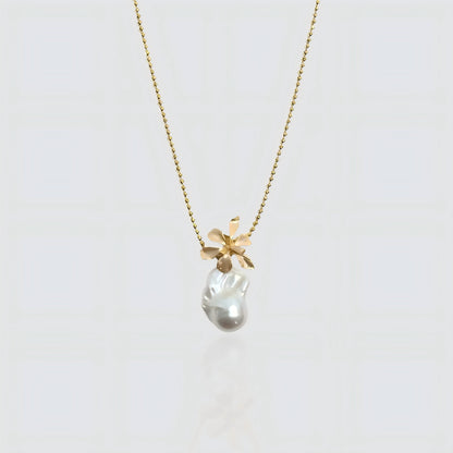 A white freshwater baroque pearl pendant topped with a sculpted gold flower design, hanging on a gold beaded satellite chain against a white background.