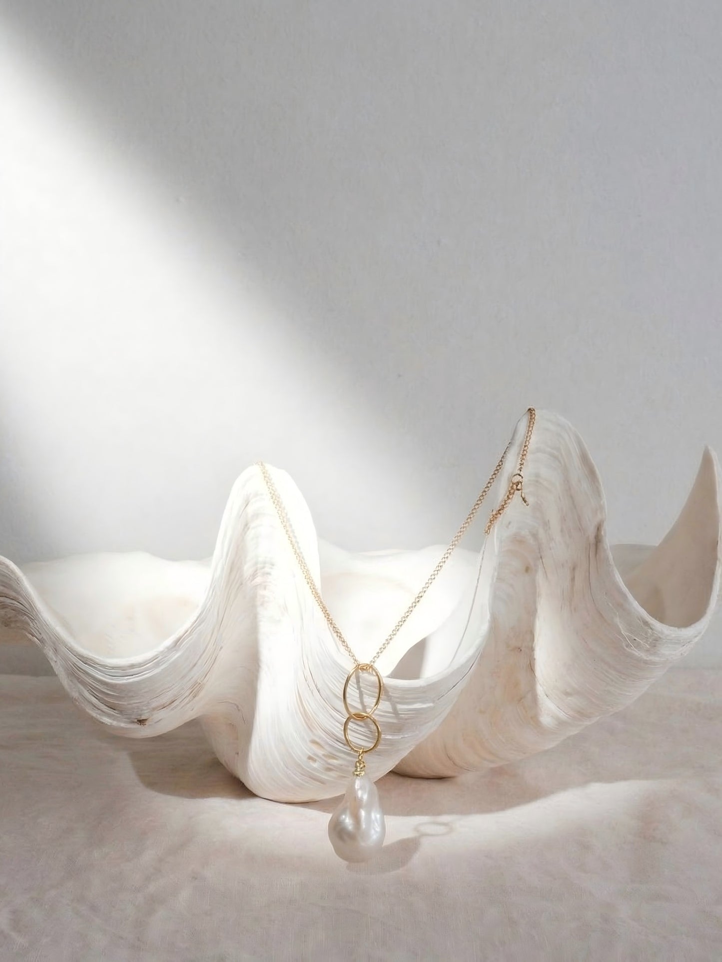 Aesthetic still life of a 14k gold interlocking ring necklace with a natural baroque pearl drop, delicately draped inside a large, sculpted white seashell.