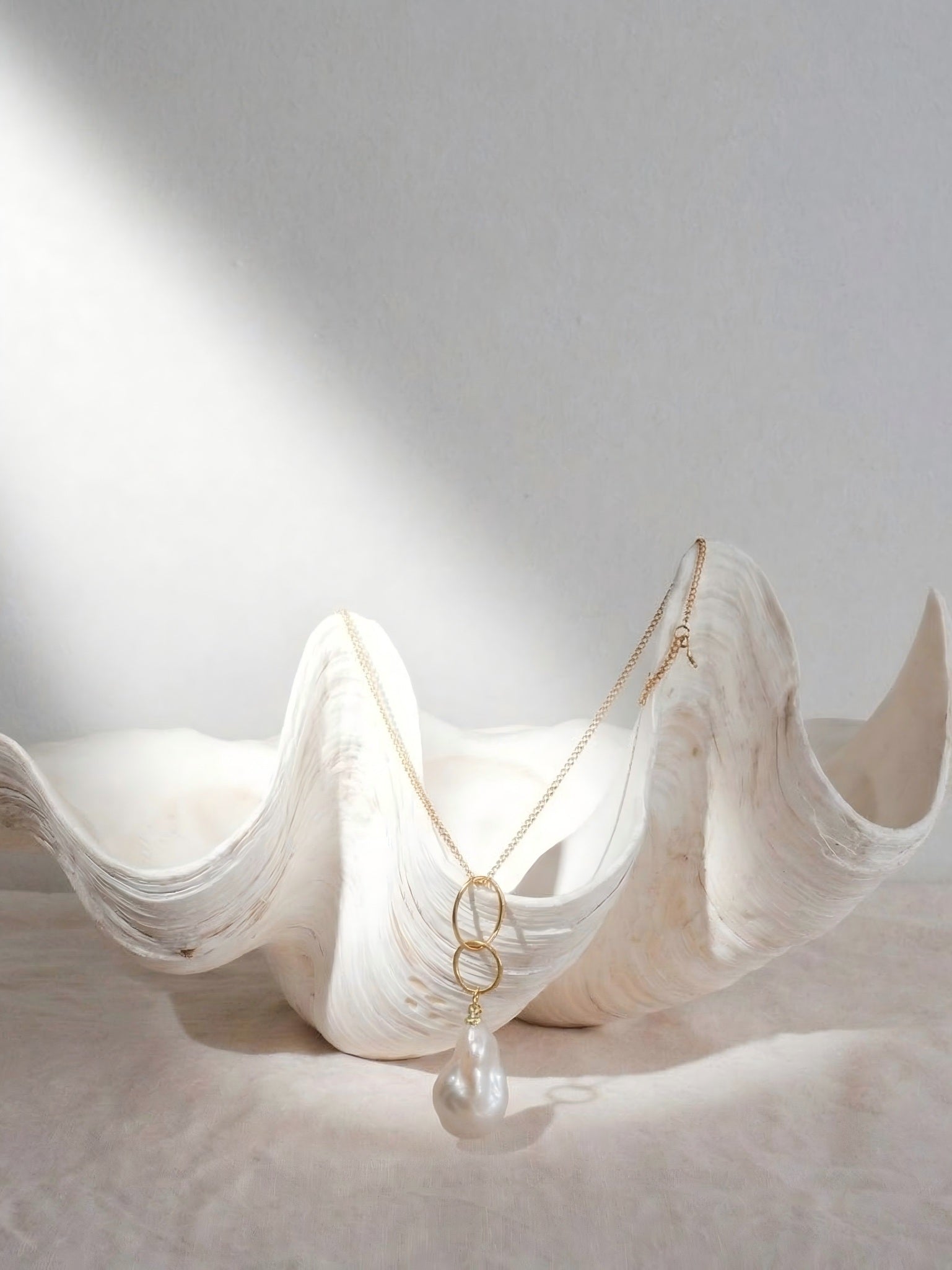 Aesthetic still life of a 14k gold interlocking ring necklace with a natural baroque pearl drop, delicately draped inside a large, sculpted white seashell.