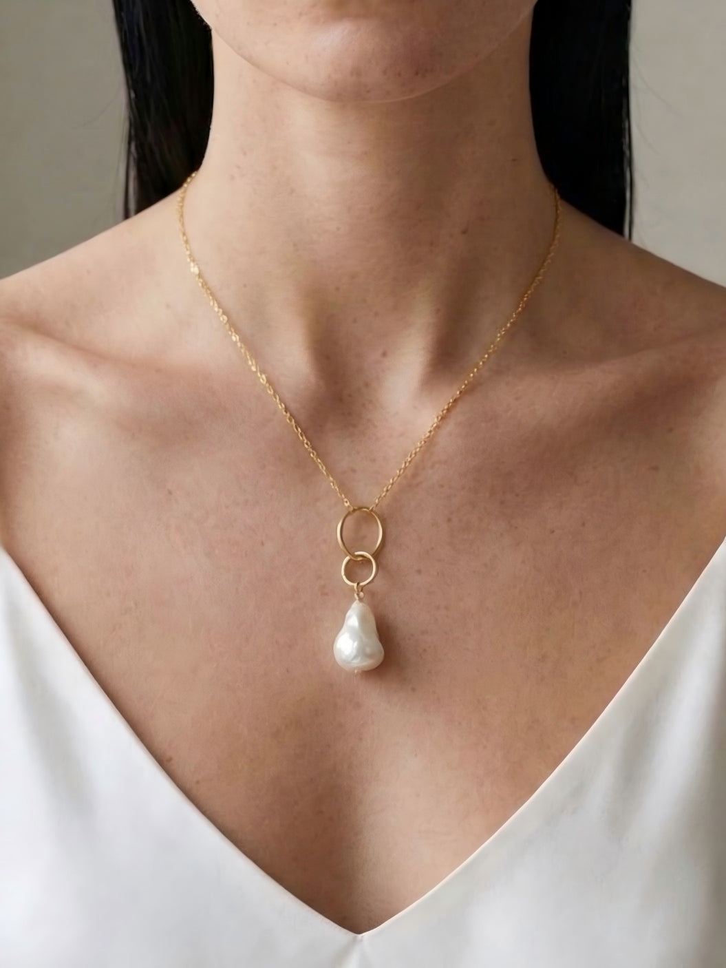 Close-up of a woman wearing a delicate 14k gold necklace with an interlocking circle pendant and a natural baroque pearl drop, resting elegantly against a white v-neck top.