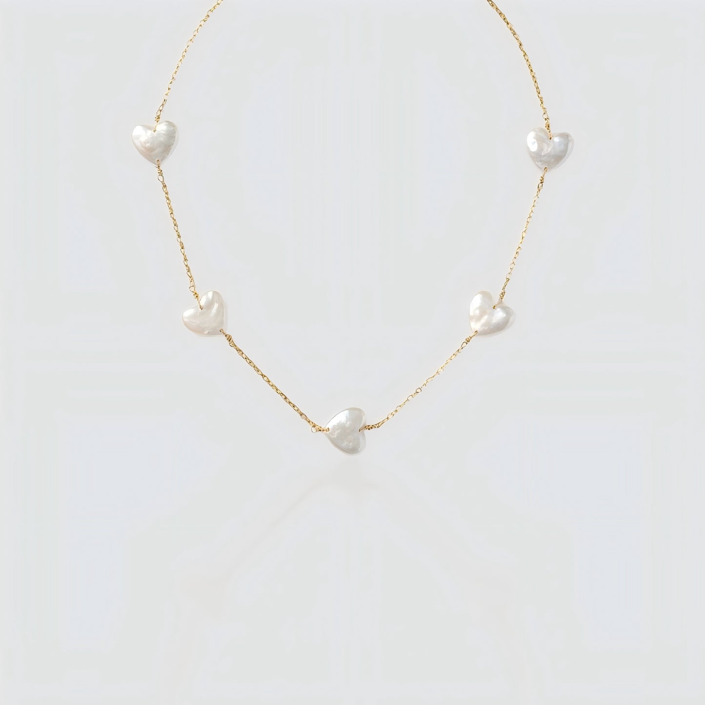 Studio shot of a gold chain necklace featuring multiple flat white heart-shaped pearls spaced out on a grey background.