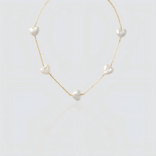 Studio shot of a gold chain necklace featuring multiple flat white heart-shaped pearls spaced out on a grey background.