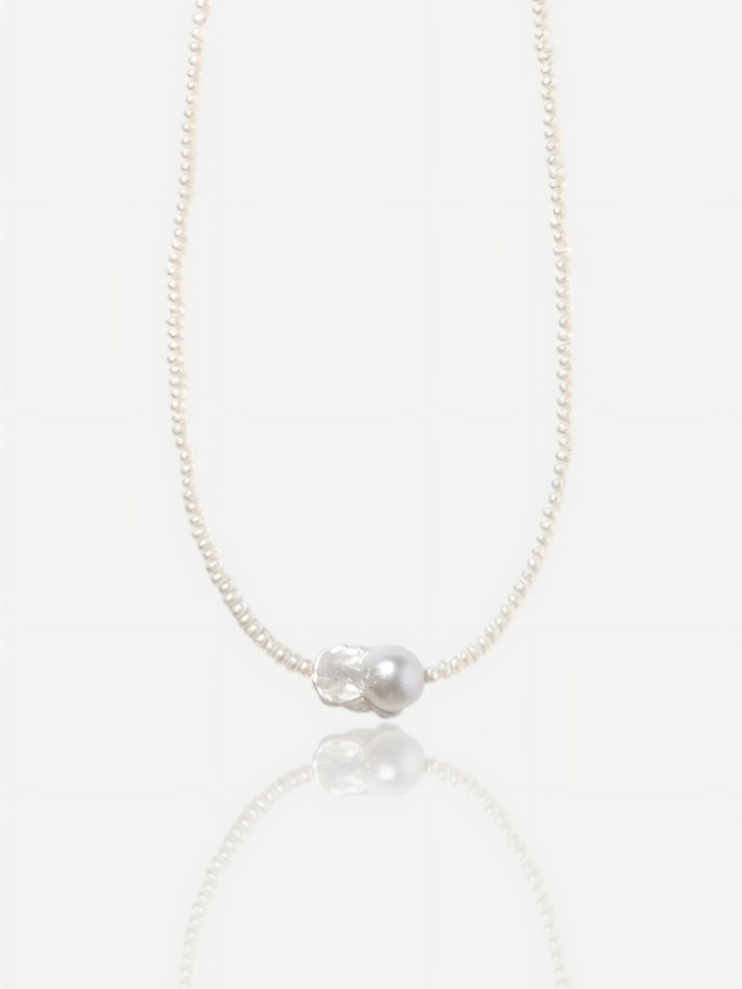 Close-up of Pearltide Azure Aura necklace featuring a large textured "crushed silver" baroque pearl centerpiece surrounded by delicate tiny seed pearls on a white background with a soft reflection, elegant quiet luxury jewelry.