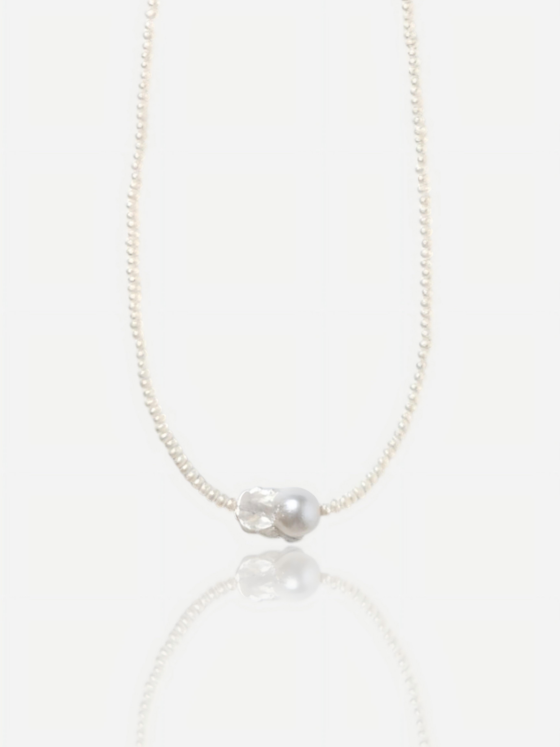 Close-up of Pearltide Azure Aura necklace featuring a large textured "crushed silver" baroque pearl centerpiece surrounded by delicate tiny seed pearls on a white background with a soft reflection, elegant quiet luxury jewelry.