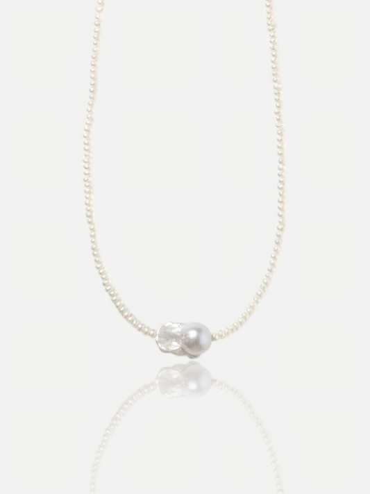 Close-up of Pearltide Azure Aura necklace featuring a large textured "crushed silver" baroque pearl centerpiece surrounded by delicate tiny seed pearls on a white background with a soft reflection, elegant quiet luxury jewelry.