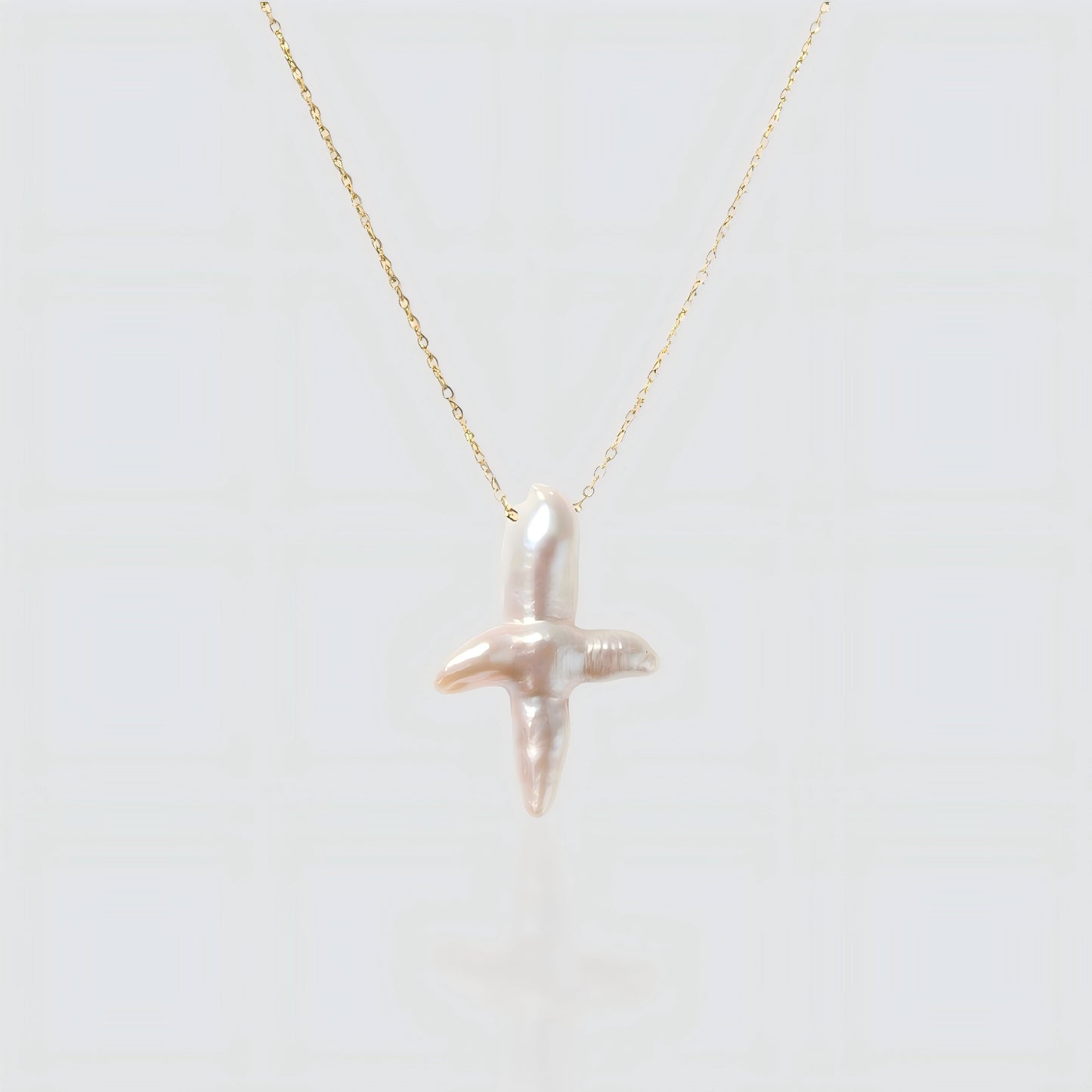 A white background product shot of a unique cross-shaped baroque pearl pendant on a delicate gold chain.