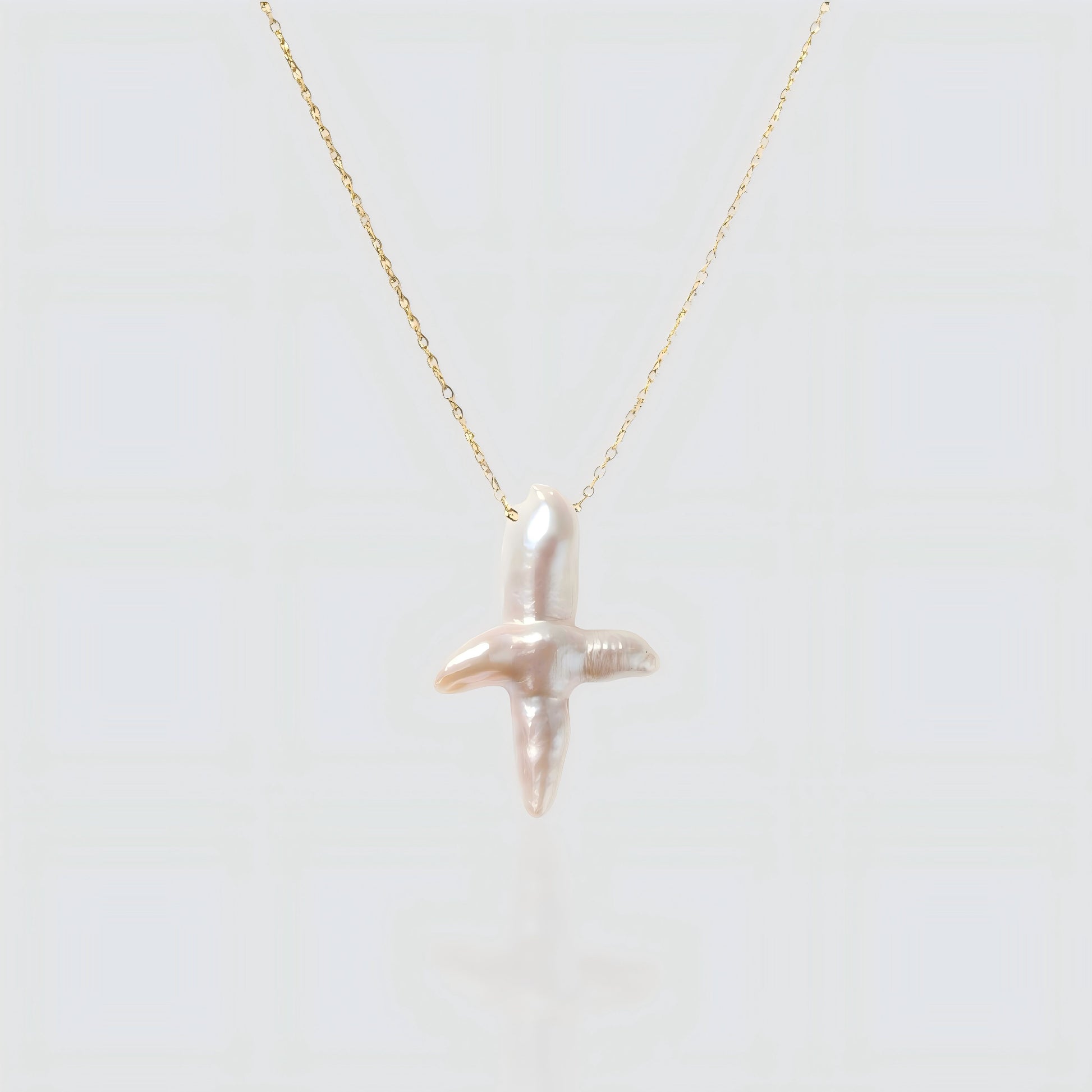 A white background product shot of a unique cross-shaped baroque pearl pendant on a delicate gold chain.