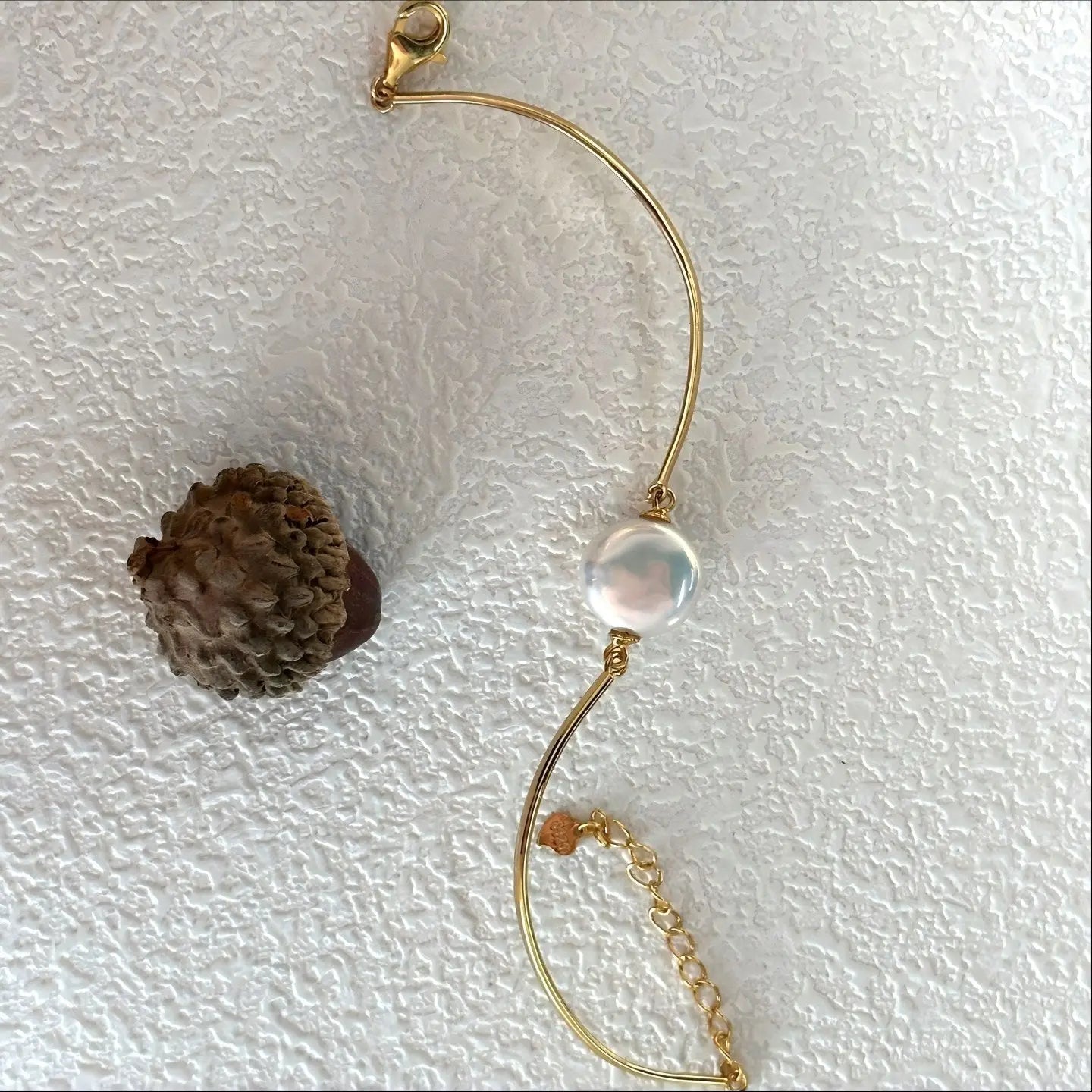 Macro shot of the center of the gold bracelet, focusing on the connection between the gold wire and the round white pearl. The image highlights the smooth surface and reflection of the pearl.