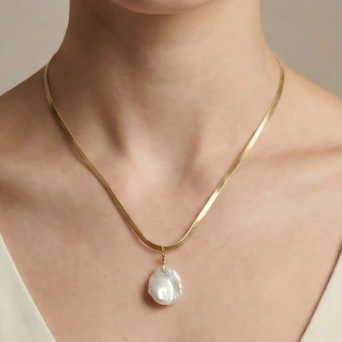 A close-up of a woman's collarbone wearing the flat gold snake chain necklace with a pearl drop. The image shows how the chain lays flat against the skin, reflecting light, while the baroque pearl adds a soft, organic touch.