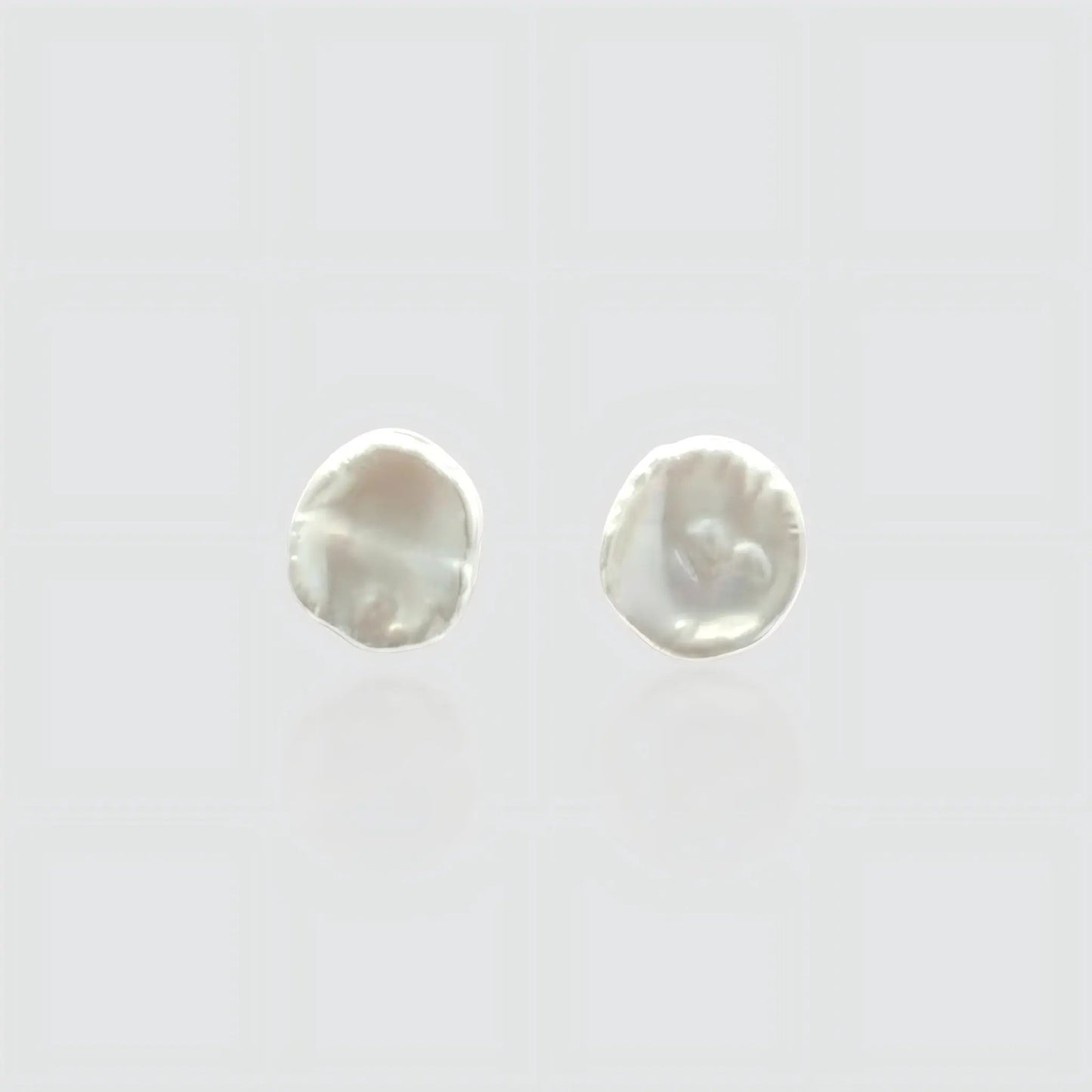 A pair of minimalist white freshwater pearl stud earrings on a light gray background. The pearls feature a flat, organic baroque shape resembling petals or coins with a high, creamy luster. Simple and elegant jewelry by Pearltide.