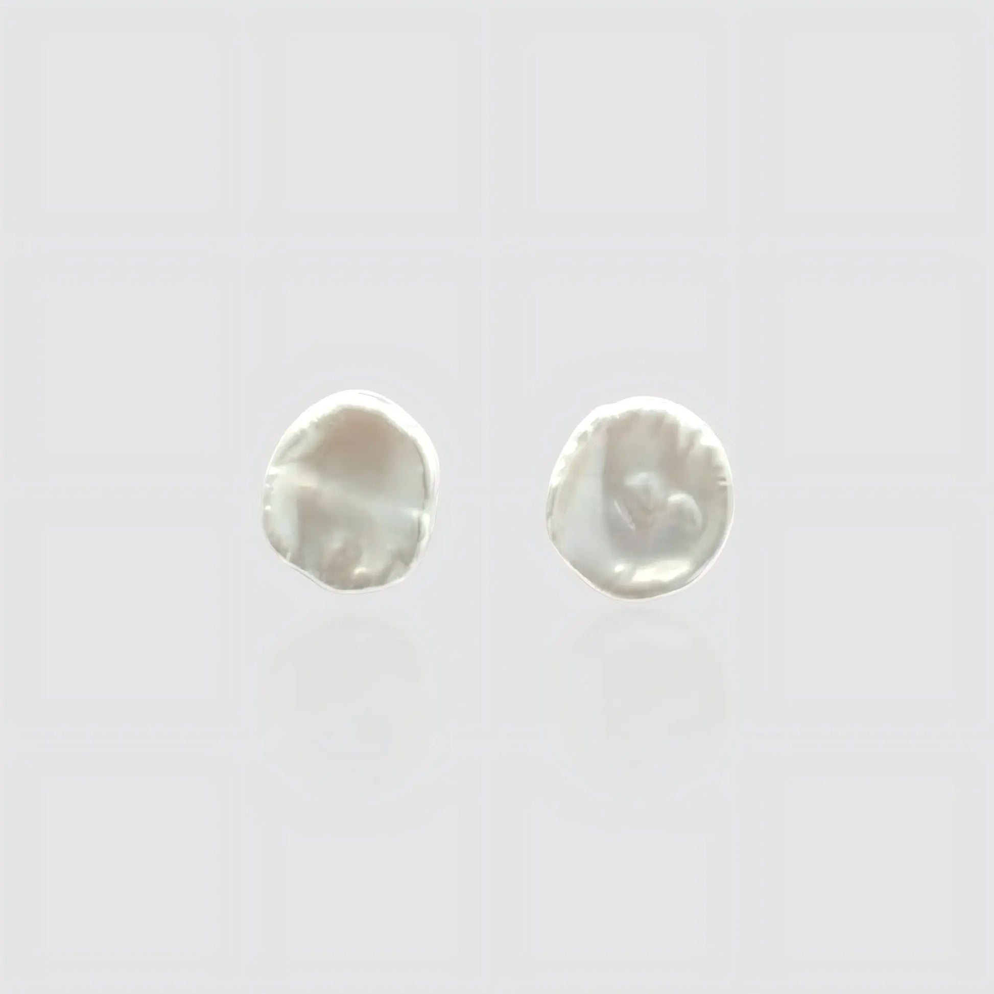A pair of minimalist white freshwater pearl stud earrings on a light gray background. The pearls feature a flat, organic baroque shape resembling petals or coins with a high, creamy luster. Simple and elegant jewelry by Pearltide.