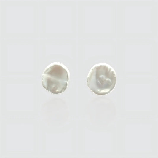 A pair of minimalist white freshwater pearl stud earrings on a light gray background. The pearls feature a flat, organic baroque shape resembling petals or coins with a high, creamy luster. Simple and elegant jewelry by Pearltide.