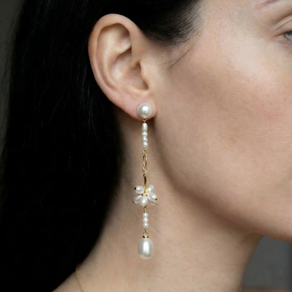 A model with styled hair wearing the Pearltide floral pearl drop earrings. The image shows the earrings dangling elegantly against the side of her neck, illustrating their long length and how the pearl luster catches the light. Perfect as bridal earrings or for formal events.