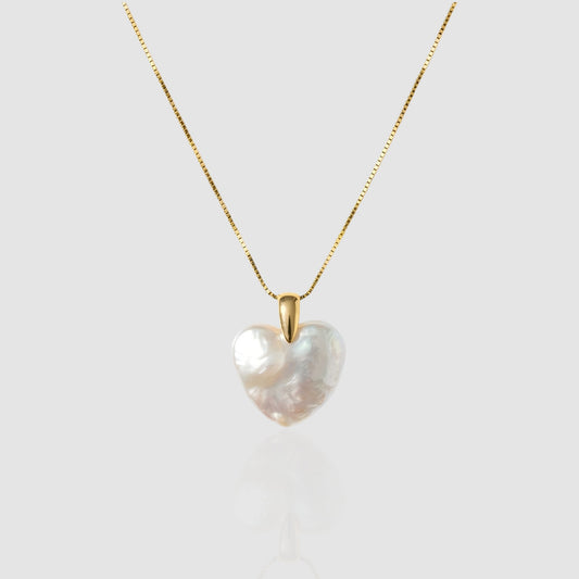 A lustrous, organic heart-shaped baroque pearl pendant hanging on a gold box chain against a light grey/white background.