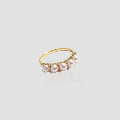 Front view of a gold band ring featuring five round pink pearls and small sparkling diamonds on a light grey reflective surface