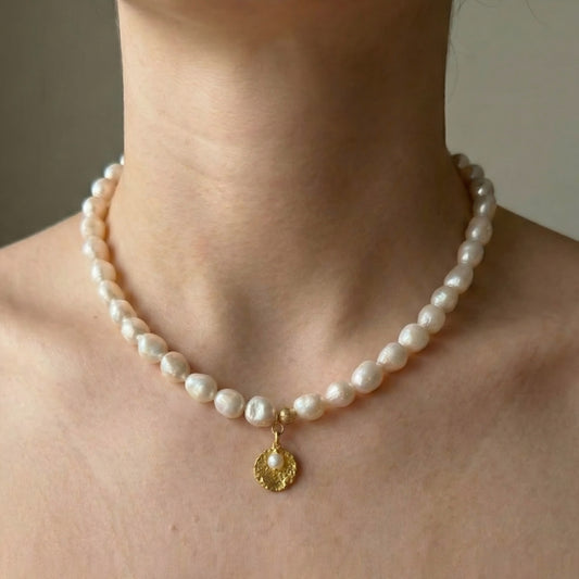 Close-up of a woman's neck wearing a classic pearl strand necklace with a hammered gold medallion charm. Romantic and elegant bridal or daily jewelry style.