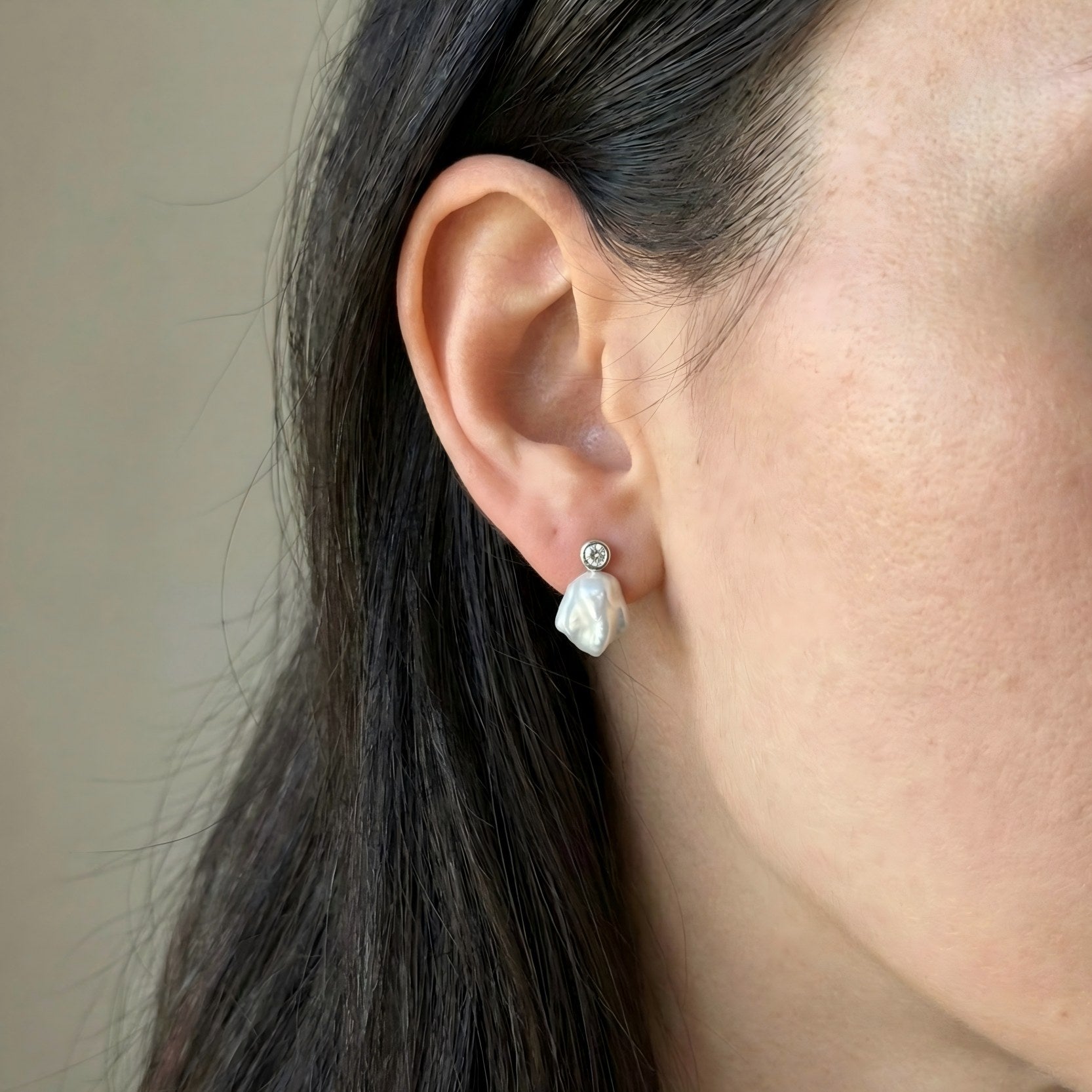 Side profile of a woman with dark hair wearing a delicate baroque pearl drop earring with a bezel-set crystal stud.