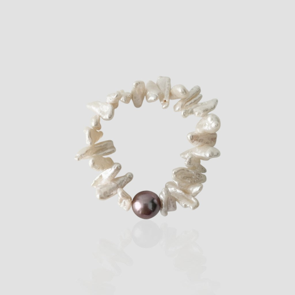 Studio shot of a sunflower-inspired pearl bracelet featuring flat white keshi pearls resembling petals and a single dark purple metallic pearl on a light grey background.