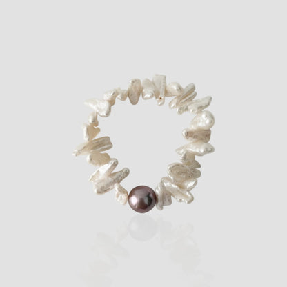 Studio shot of a sunflower-inspired pearl bracelet featuring flat white keshi pearls resembling petals and a single dark purple metallic pearl on a light grey background.