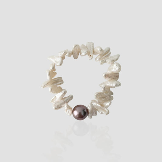 Studio shot of a sunflower-inspired pearl bracelet featuring flat white keshi pearls resembling petals and a single dark purple metallic pearl on a light grey background.